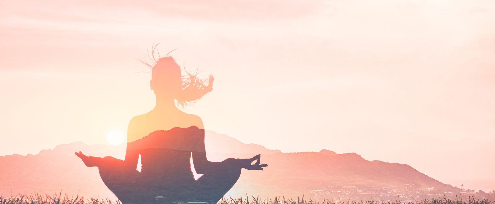 A woman seated in meditation against a sunrise over the hills